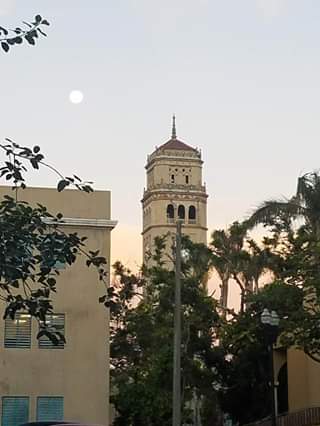Foto de la Torre de la Universidad de Puerto Rico (UPR), Recinto de Río Piedras, en un amanecer con la luna de fondo en el cielo.